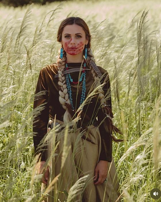 Lakota Gordon standing in a field of tall grass wearing traditional jewelry and a red handprint, in honor of Missing and Murdered Indigenous Women